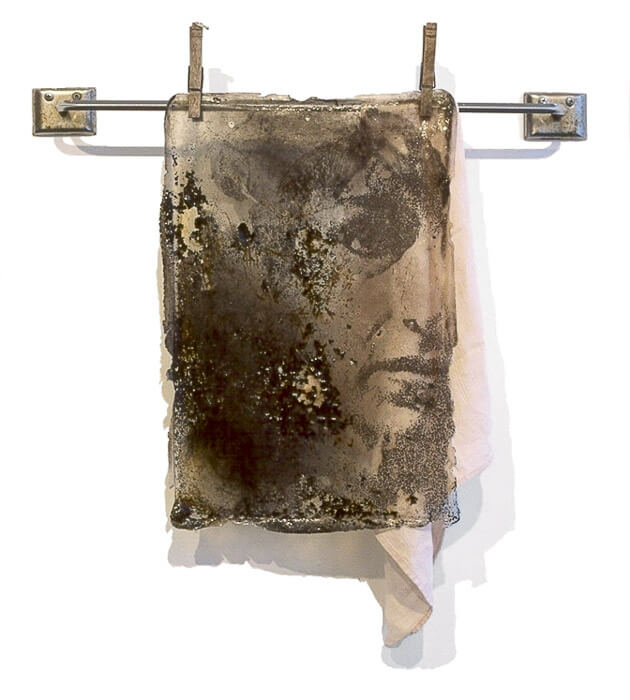 artwork with a photographic image transfer of a woman's face behind rust and carbon embedded in acrylic and hung on a rack with a napkin behind the face