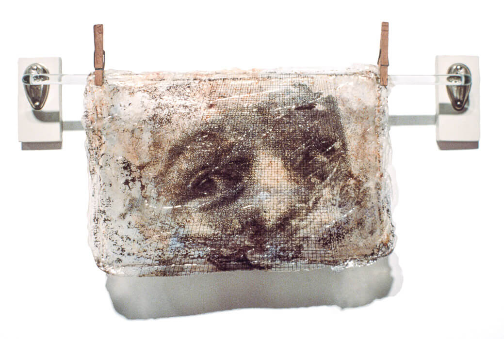 A mixed media artwork with eyes looking through a grid on a translucent acrylic skin with rust and carbon, hung with found objects.